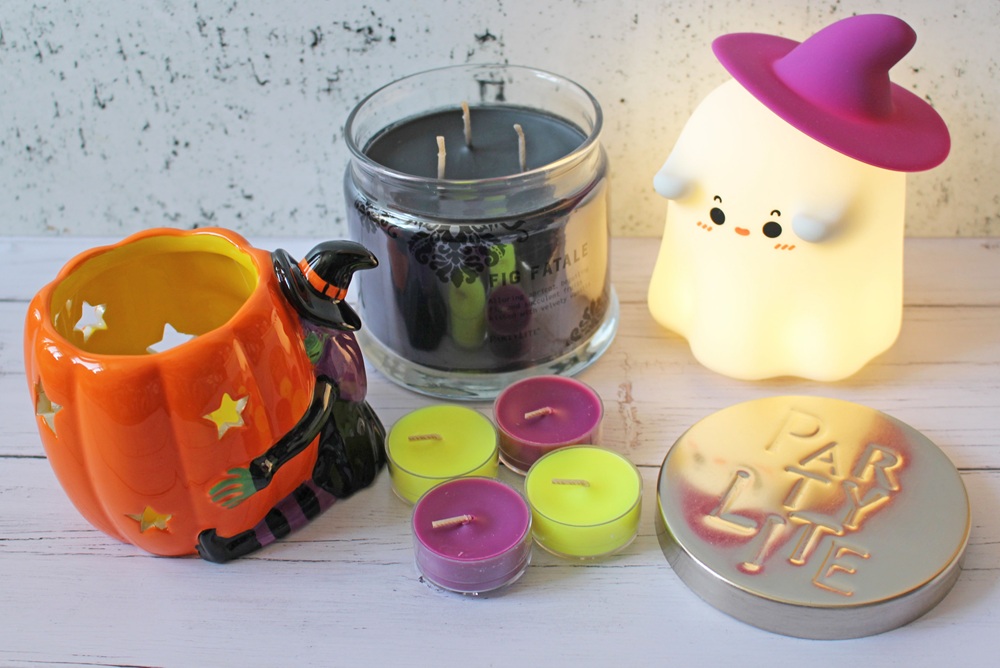 A Hallowe’en-themed display with a pumpkin-shaped candle holder, a witch figurine, a glowing ghost lamp with a pink hat, a black candle, four small tealights, and a tin labelled PARTYLITE on a white surface.