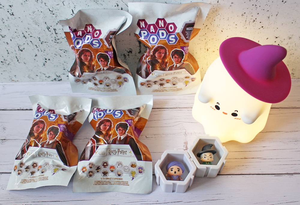 Four Harry Potter-themed Nano Pods toy packets are displayed on a white surface beside a cute ghost-shaped lamp with a purple witch’s hat. Two opened hexagonal pods reveal mini figures of Harry Potter and Hermione Granger.