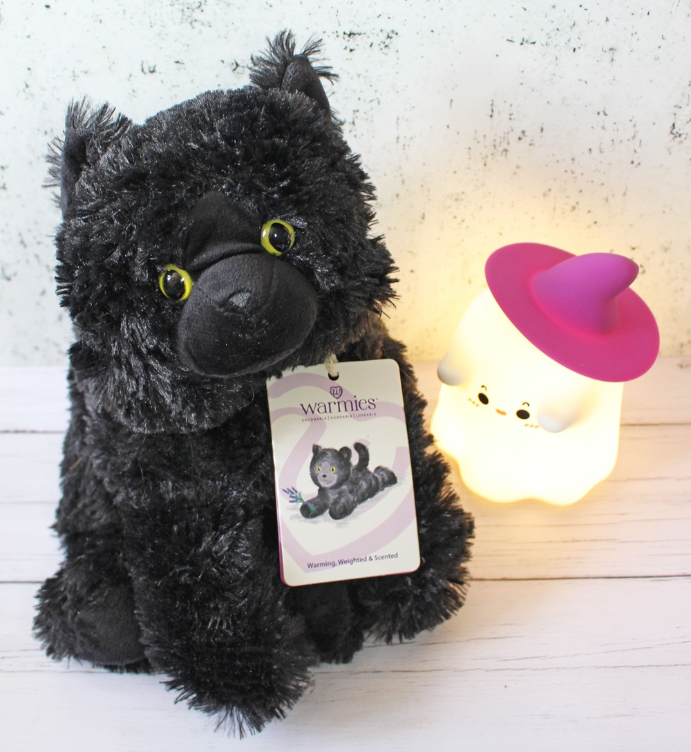 A plush black cat with yellow-green eyes and a Warmies tag sits next to a glowing ghost-shaped night light with a pink witch's hat on a white wooden surface against a speckled wall.