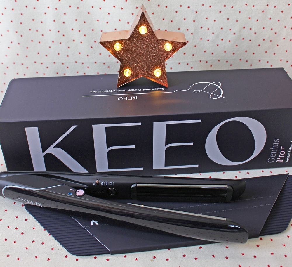 A black KEEO Genius Pro+ hair straightener rests on its box, with a lit star-shaped decoration in the background and a white fabric with small red polka dots underneath.