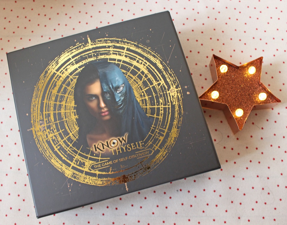 A box for Know Thyself: The Game of Self-Discovery featuring half a human face and half a masked face, with a gold circular design, sits next to a lit star-shaped decoration on a polka-dotted surface.