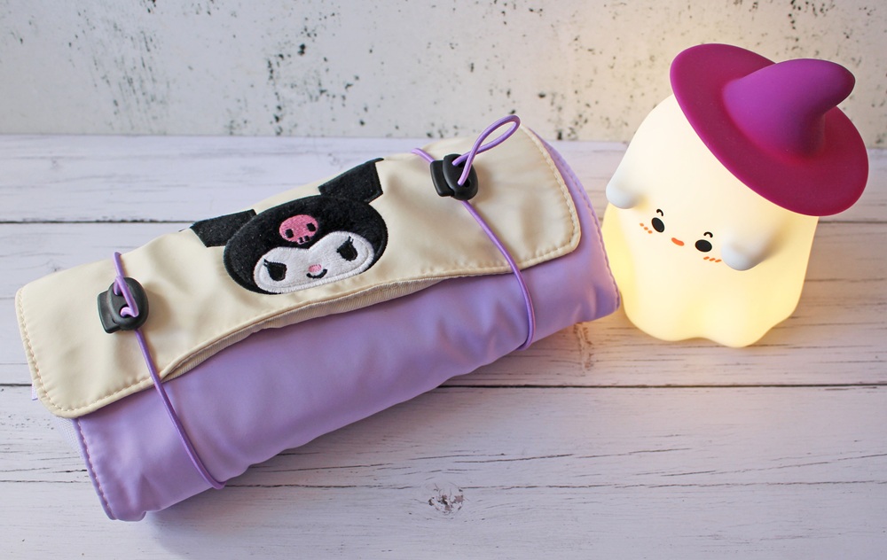 A rolled-up, pastel purple bag with a Kuromi character design next to a cute, glowing ghost lamp with a purple witch hat on a white wooden surface.