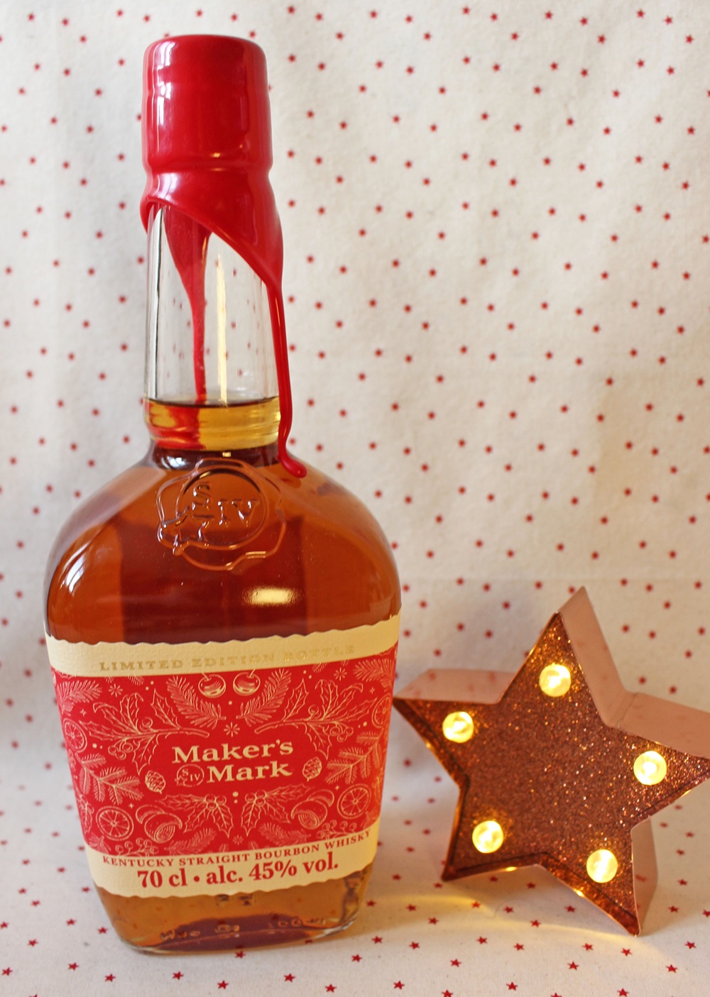 A bottle of Maker’s Mark bourbon with a red wax seal stands next to a lit, glittery star decoration, against a white background with small red polka dots.