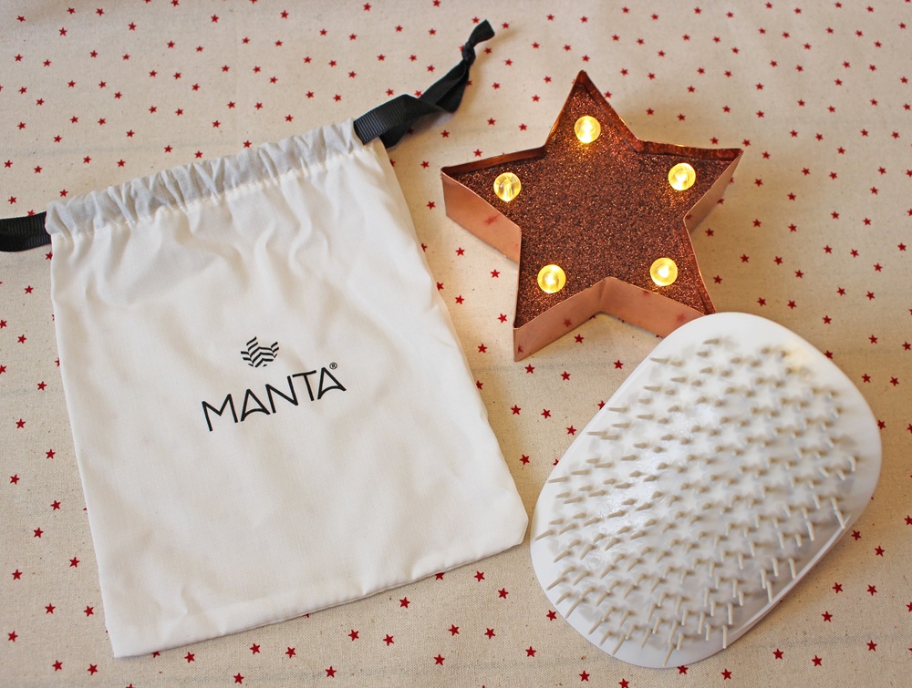 A white MANTA drawstring bag, a white hairbrush with flexible bristles, and a lit, star-shaped decorative light are arranged on a beige cloth with small red stars.