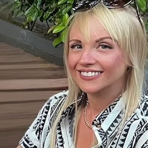 A smiling woman with straight blonde hair, wearing sunglasses on her head and a patterned shirt, sits outdoors near wooden steps and greenery.