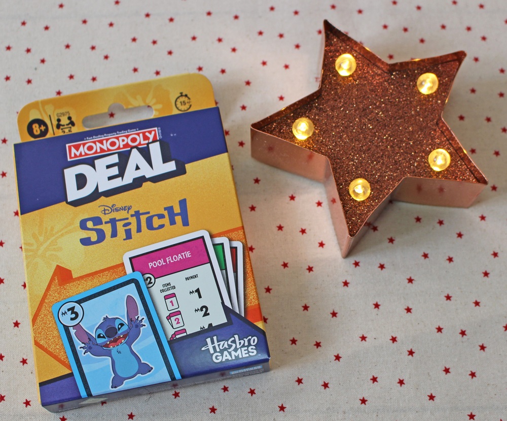 A Monopoly Deal Disney Stitch card game sits on a white background with red stars, next to a lit, glittery star-shaped ornament.