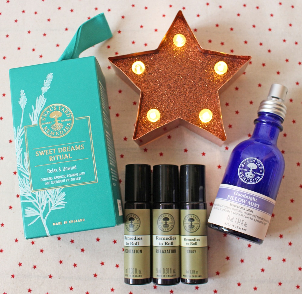 Gift set with Neal's Yard Remedies products, including Sweet Dreams Ritual box, three roll-on oils (Study, Relaxation, Night Time), a blue bottle of Goodnight Pillow Mist, and a lit star decoration on a red star-patterned cloth.