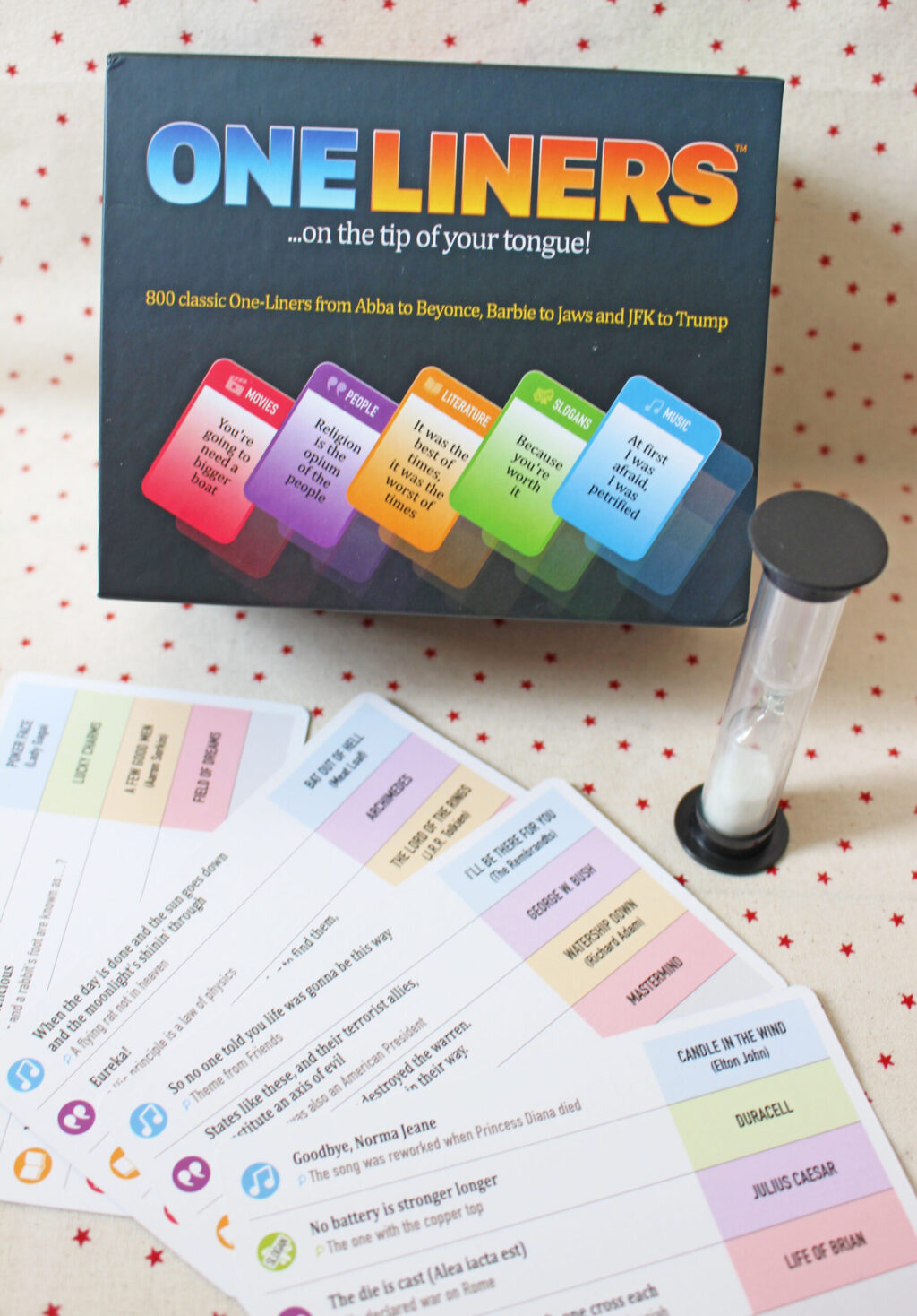 A board game called One Liners is shown with its box, several colourful cards spread out, and a small hourglass timer, all placed on a white surface with red dots.