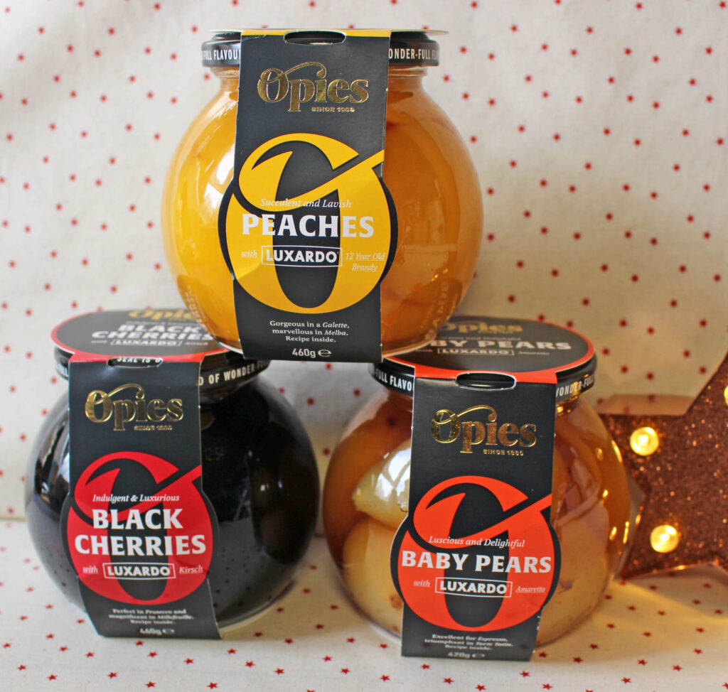Three glass jars of Opies fruit in syrup are stacked: peaches, black cherries, and baby pears, each labelled with Luxardo. A lit star decoration is partially visible in the background.