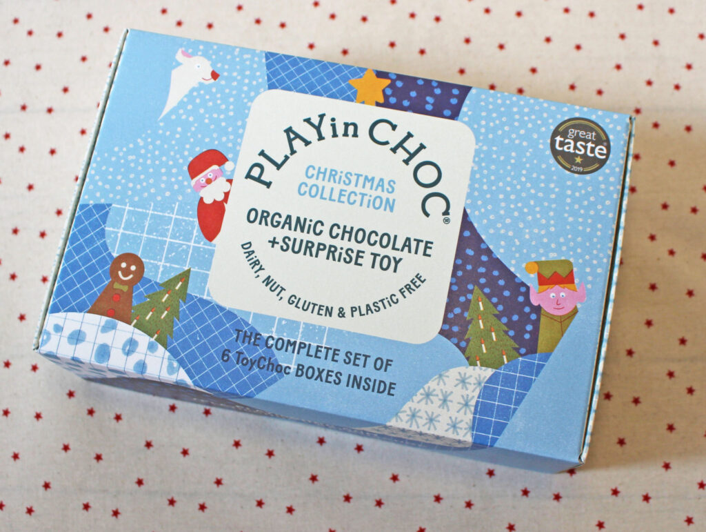 A blue box labelled PLAYin CHOC Christmas Collection with festive illustrations, advertising organic chocolate and a surprise toy that is dairy, nut, gluten, and plastic free. The box is on a cream fabric with red dots.