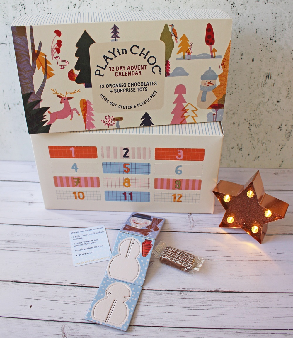 A PLAYin CHOC 12-day advent calendar box stands open, revealing numbered doors. Beside it are a glowing star-shaped light, a chocolate treat, a paper bunny cut-out, and a small note, all on a white wooden surface.