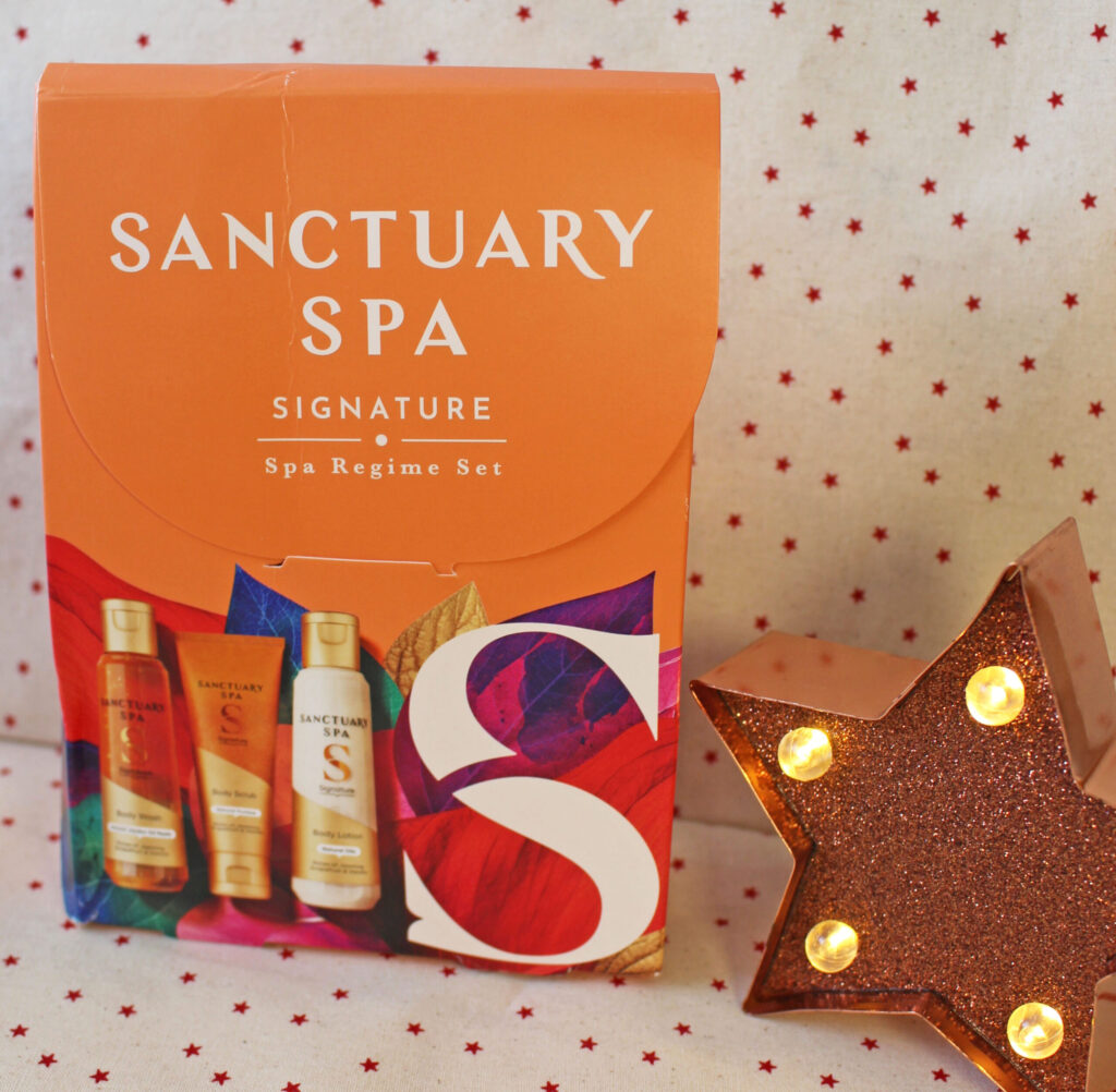 An orange Sanctuary Spa Signature Spa Regime Set box with colourful accents sits next to a lit, star-shaped decoration on a white background with small red stars.