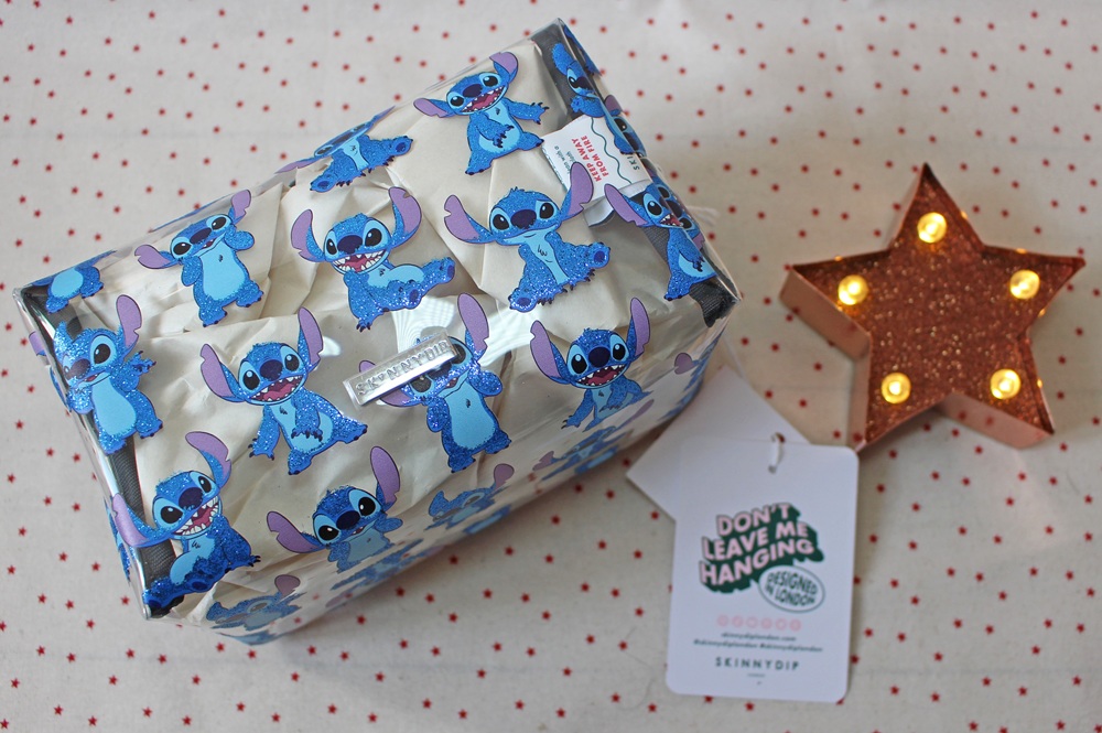 A rectangular pouch decorated with blue Stitch characters from Lilo & Stitch sits on a spotty surface beside a lit gold star ornament and a tag that reads DON'T LEAVE ME HANGING.