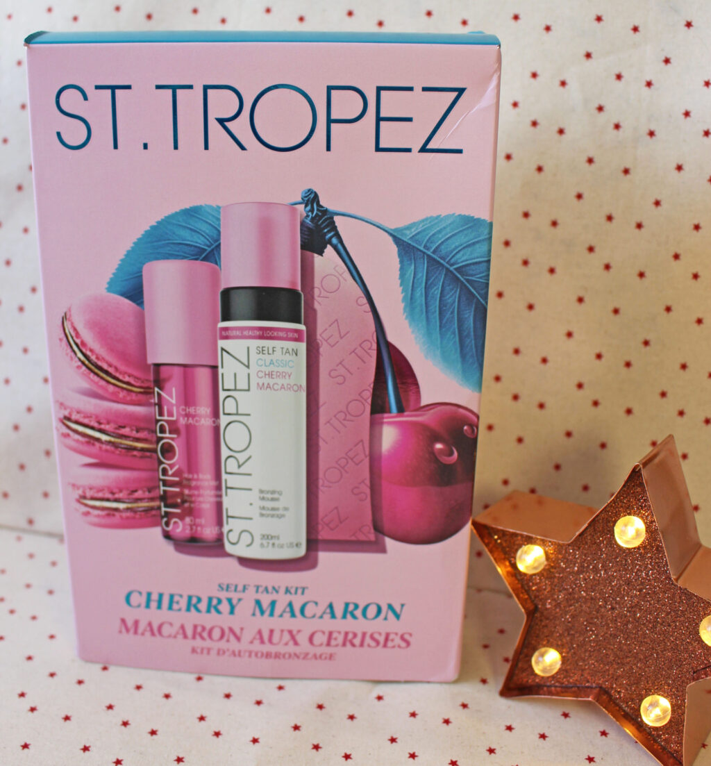 A St. Tropez Cherry Macaron self-tan kit box featuring two self-tan products against a pink backdrop with cherry and macaron illustrations. A lit star-shaped decoration is visible in the lower right corner.