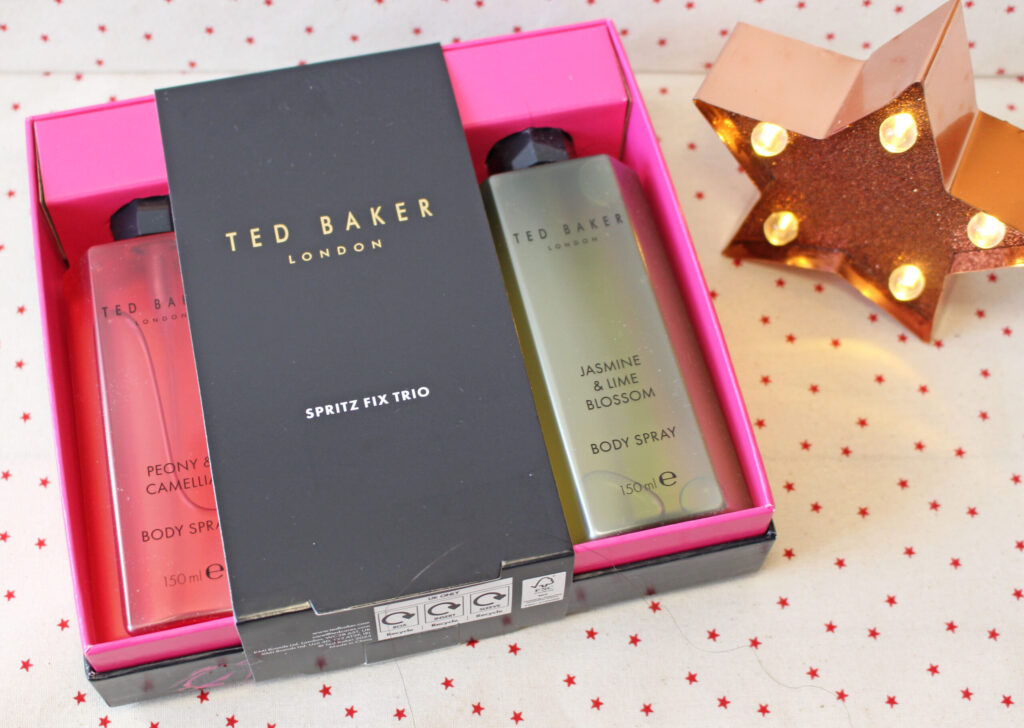 A Ted Baker London Spritz Fix Trio gift set with two body sprays—Peony & Camellia and Jasmine & Lime Blossom—packaged in a black and pink box, next to a lit, star-shaped light on a star-patterned surface.