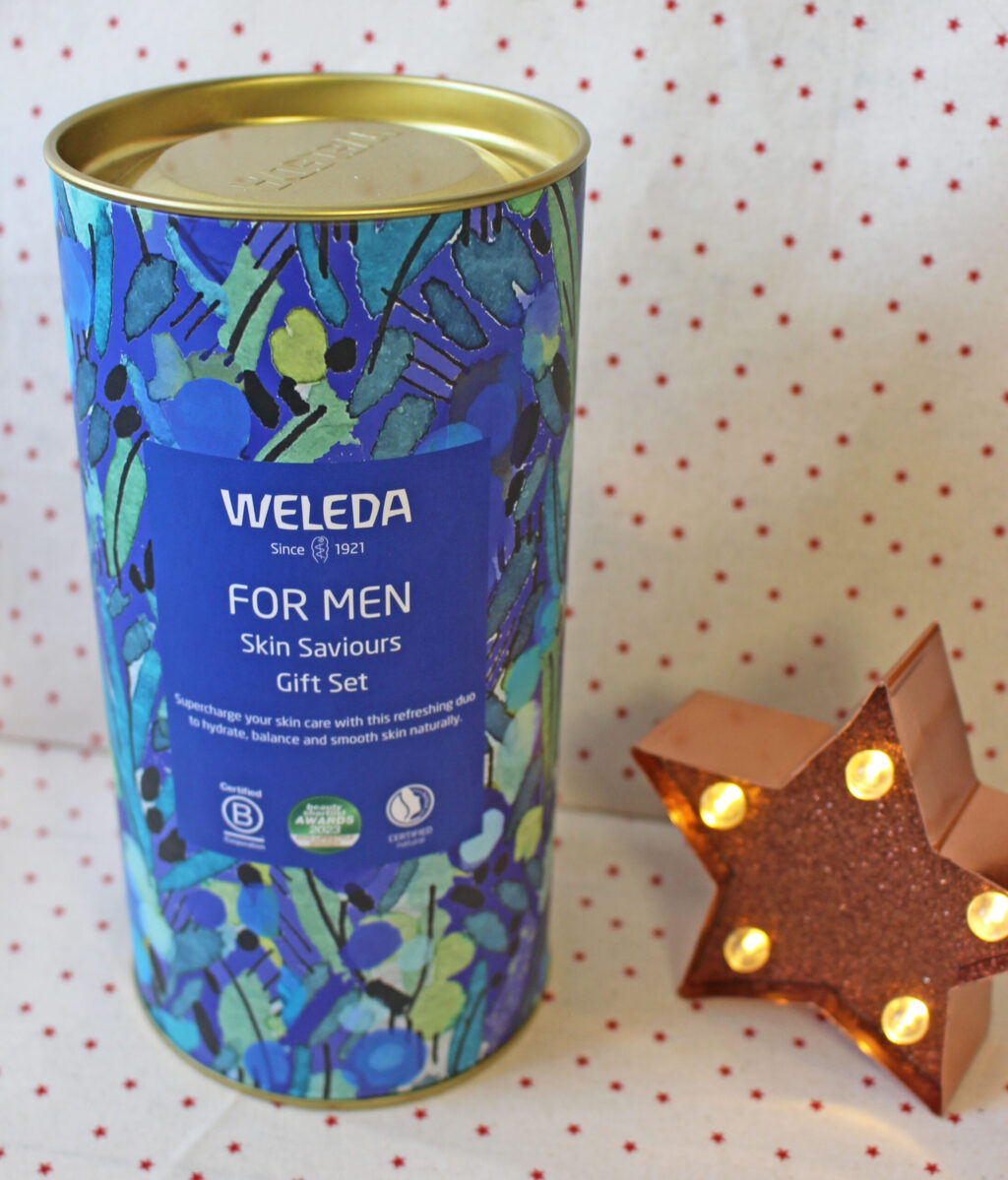 A cylindrical Weleda For Men Skin Saviours gift set with a blue and green floral design stands next to a lit, brown star-shaped ornament on a white surface with red dots.