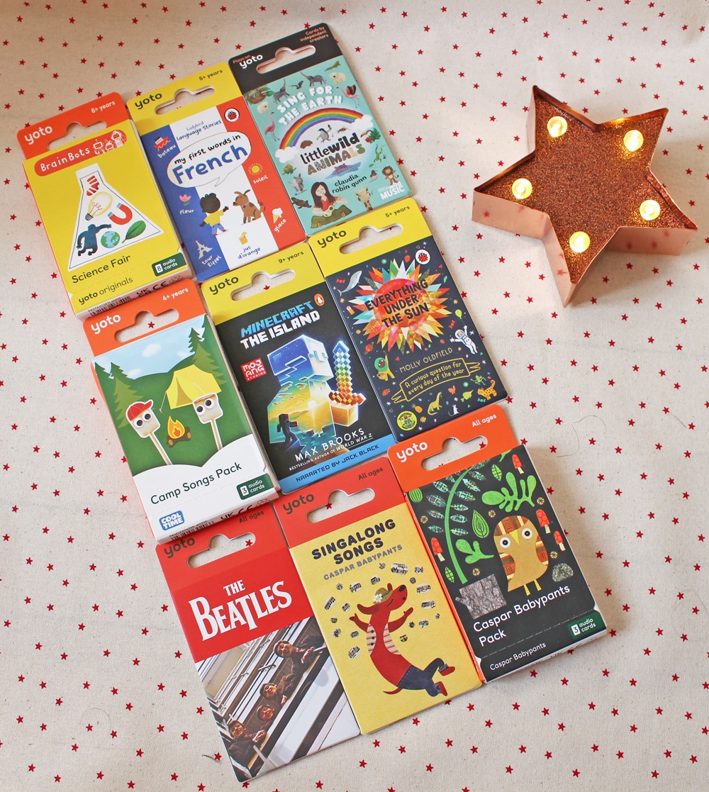 Nine colourful Yoto card packs are arranged in a grid on a fabric with red stars, next to a lit star-shaped lamp. The packs feature themes like science, languages, music, and stories.