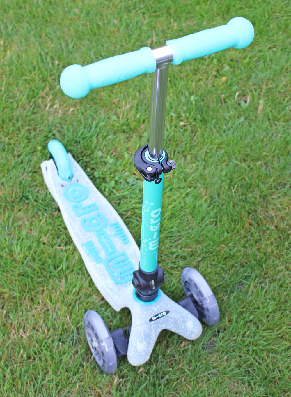 A turquoise three-wheeled children's scooter with handlebar grips and wheels is standing on green grass.