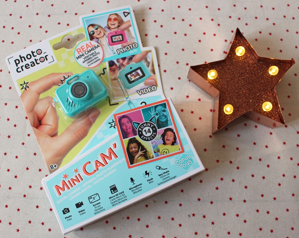A boxed “Mini Cam” toy camera kit sits on a star-patterned cloth next to a lit, copper-coloured star lamp. The box shows images of children using the mini camera for photos and videos, and has a bright, playful design.