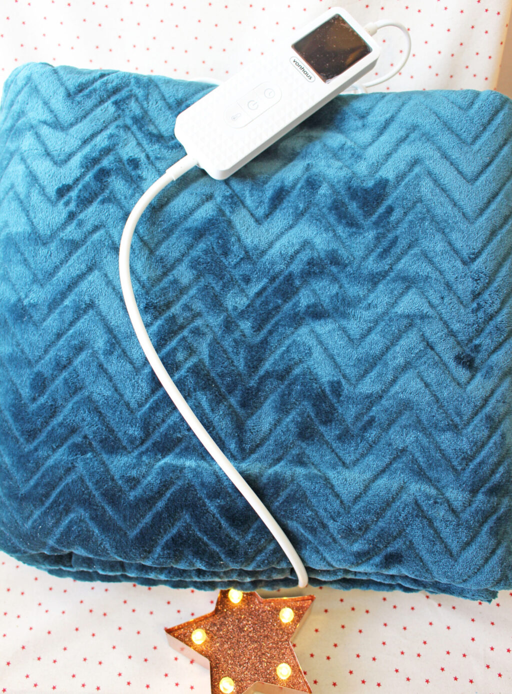 A blue textured heated blanket with a white remote control rests on a patterned surface; a lit star-shaped decoration is partially visible at the bottom.