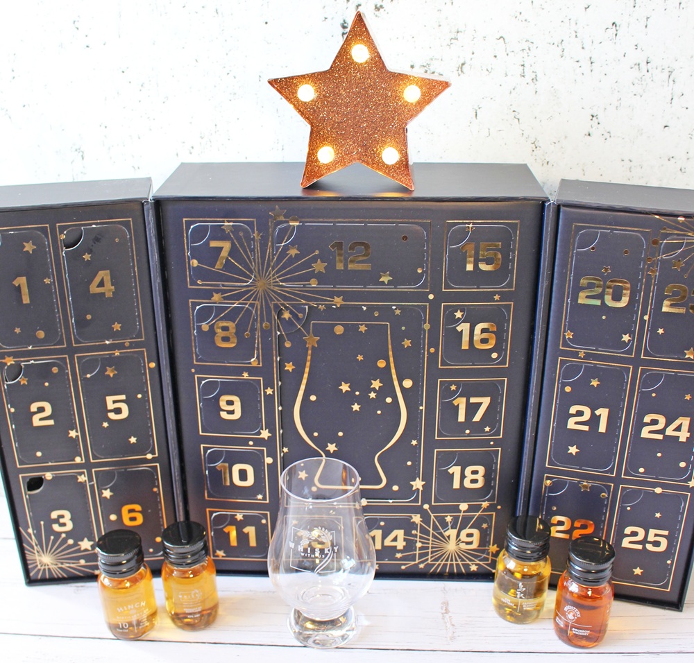 A festive advent calendar with numbered doors, a glowing star on top, a glass, and small bottles of whisky arranged in front, all set on a light wooden surface.