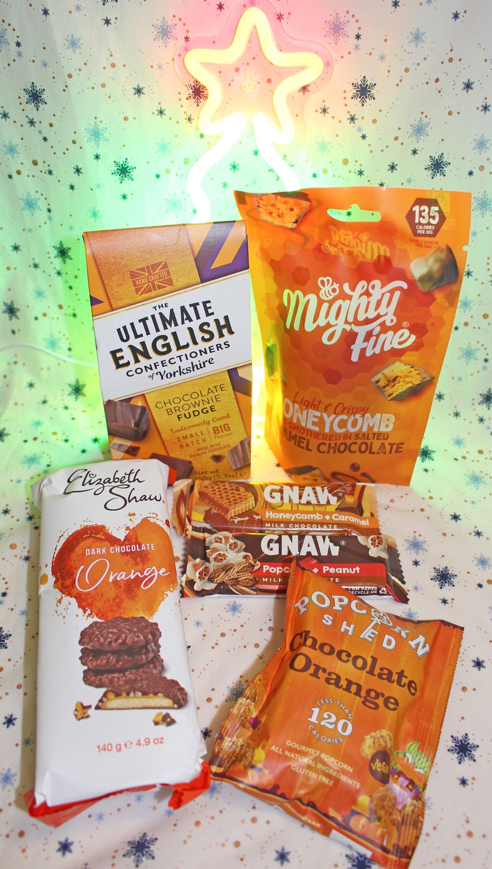 A selection of chocolate orange treats, including bars and snack packs, displayed on a white surface with blue stars, with colourful neon lights shaped like a star glowing in the background.