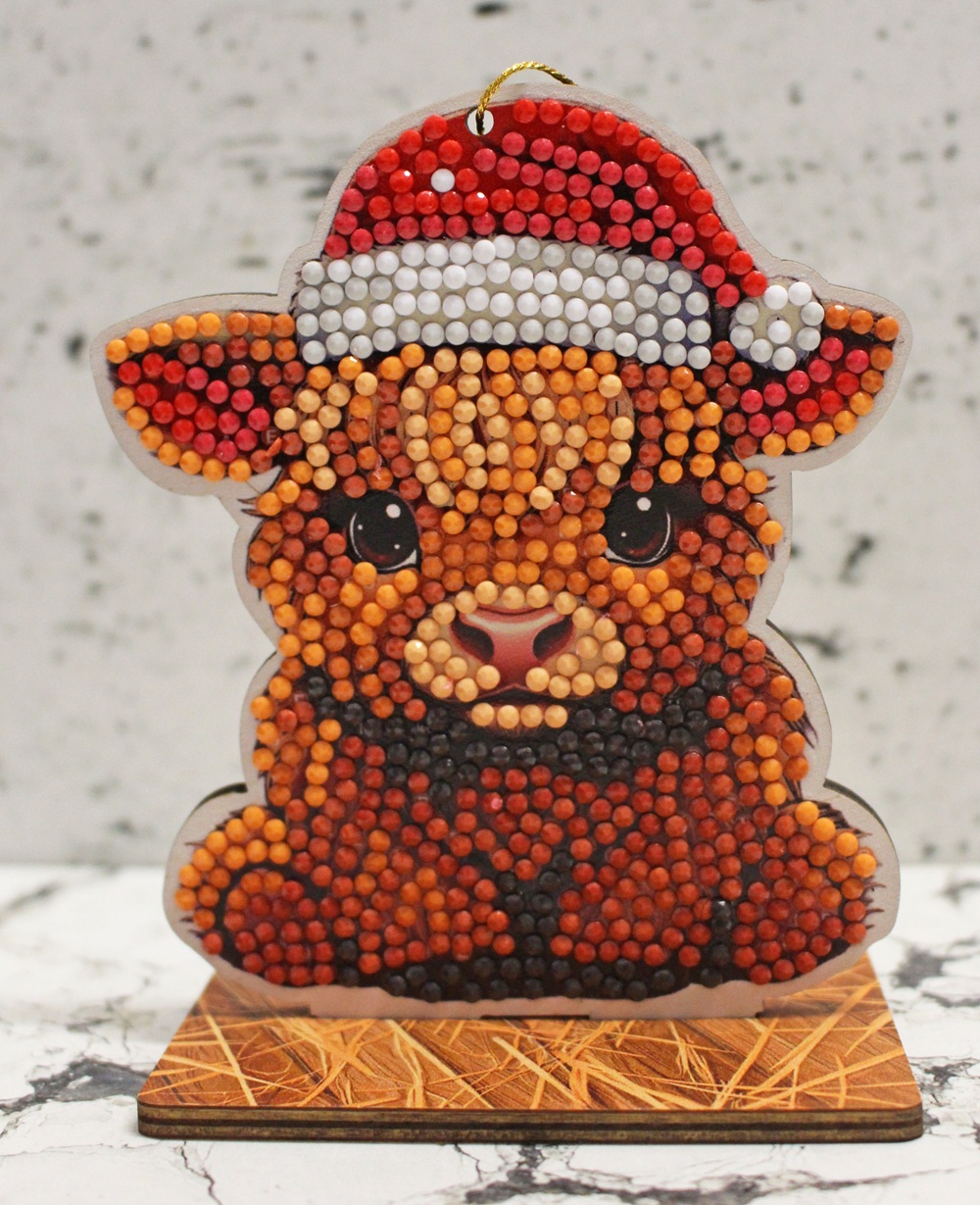 A beaded ornament of a cute brown alpaca wearing a red and white Santa hat, standing on a wooden base with a marble-patterned background, from the Craft Buddy Christmas Crystal Art collection.