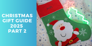 A green Christmas stocking with a Father Christmas design and the name Ash on the red cuff lies on star-patterned fabric. Text on a blue background reads: Christmas Gift Guide 2025 Part 2. Neon star and tree lights are beside the stocking.
