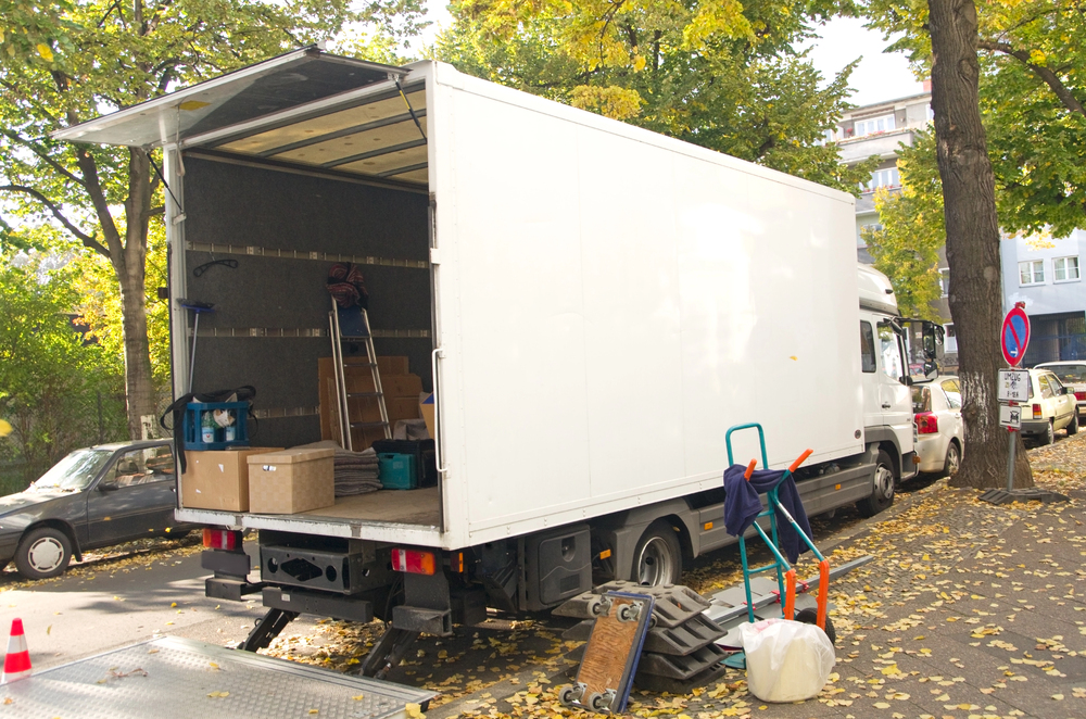 A white removal lorry is parked on a leafy street with its back door open. Boxes, a trolley, and packing materials are visible near and inside the lorry. Cars are parked along the kerb and trees with yellow leaves line the street.