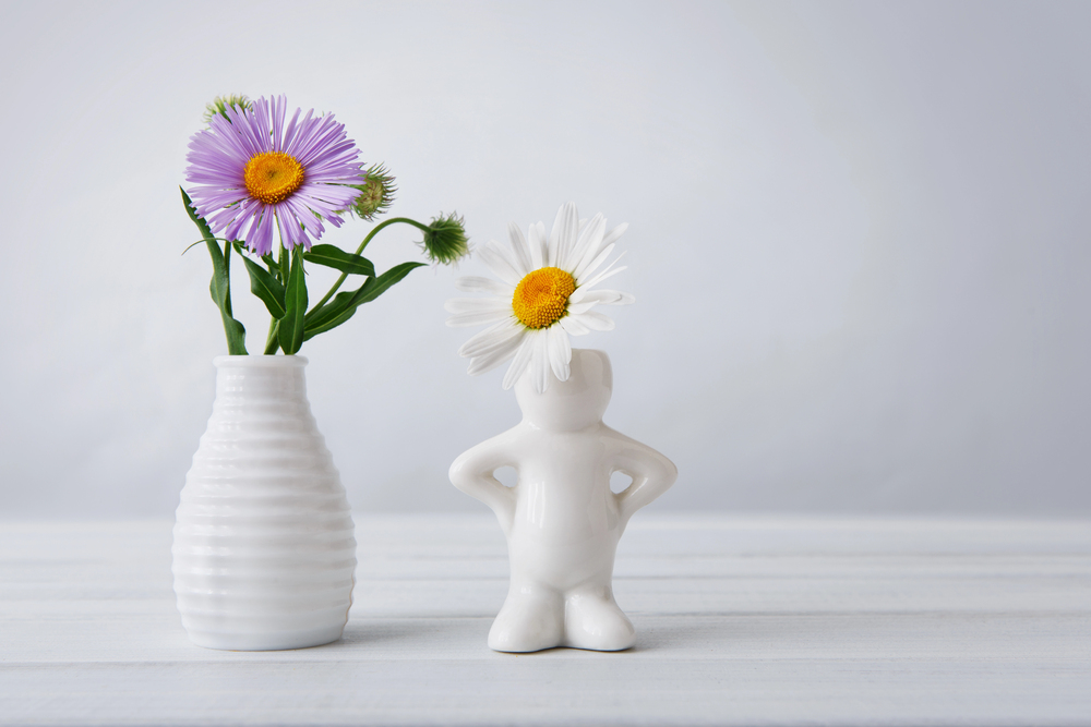 A white vase with purple and yellow flowers stands next to a white ceramic figure holding a white daisy as its head on a light wooden surface with a pale background.