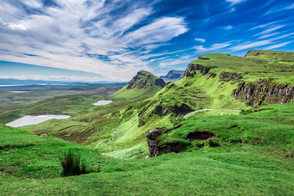 Rolling green hills and dramatic cliffs beneath a vibrant blue sky with scattered clouds. Two small lakes are visible in the distance, set within the lush landscape. The scene is bright and picturesque.