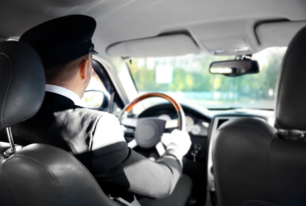 A chauffeur wearing a uniform and black cap is driving a car, viewed from the back seat. The driver is holding the steering wheel with white-gloved hands, and sunlight is streaming through the windows.