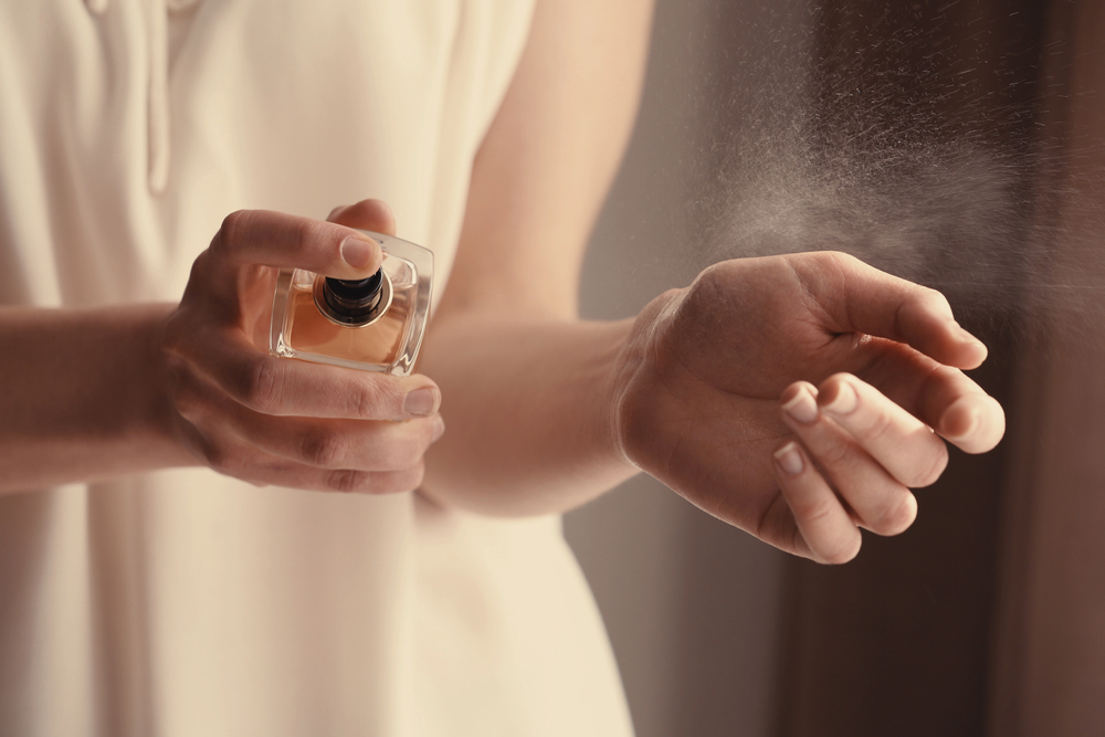 A person sprays perfume onto their wrist from a small bottle, with a fine mist visible in the air. The person is wearing a sleeveless, light-coloured top.