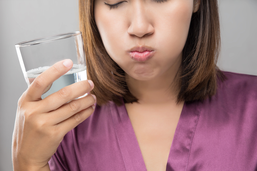 A woman in a purple top holds a glass of water in one hand and swills water in her mouth, with her cheeks puffed out. Her eyes are partially closed, and her face is partially out of frame.