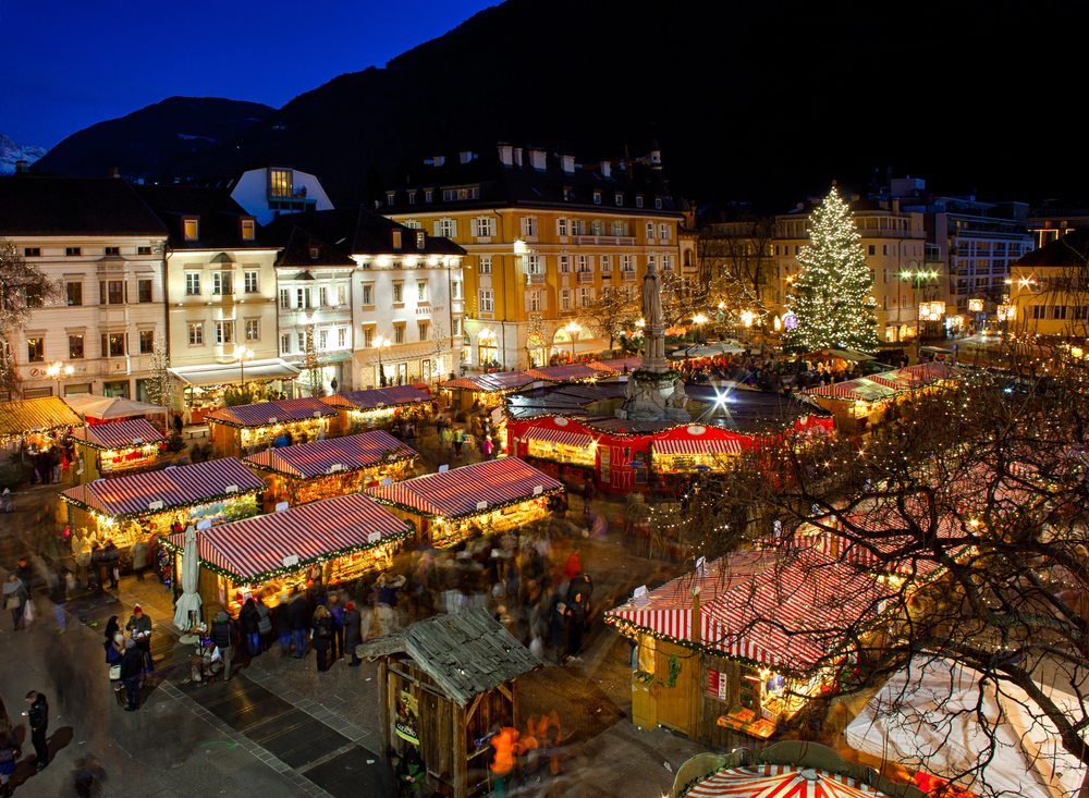 A lively outdoor Christmas market at night with red and white striped stalls, a large decorated Christmas tree, and festive lights in a town square surrounded by illuminated buildings and mountains.