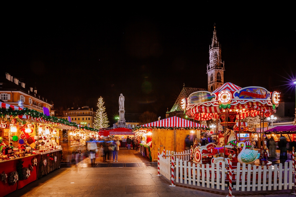 A festive Christmas market at night features colourful stalls, a brightly lit carousel, a large decorated Christmas tree, and a historic church tower under a dark sky. People walk among the lights and holiday decorations.