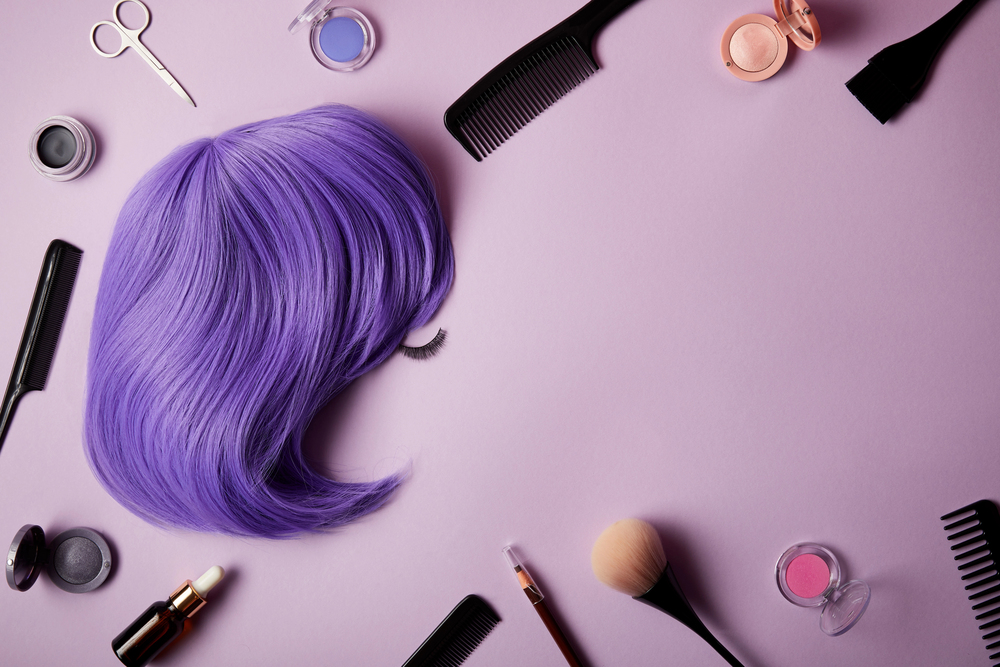 A purple wig, false eyelash, combs, make-up brushes, scissors, and cosmetics are arranged on a light purple surface.
