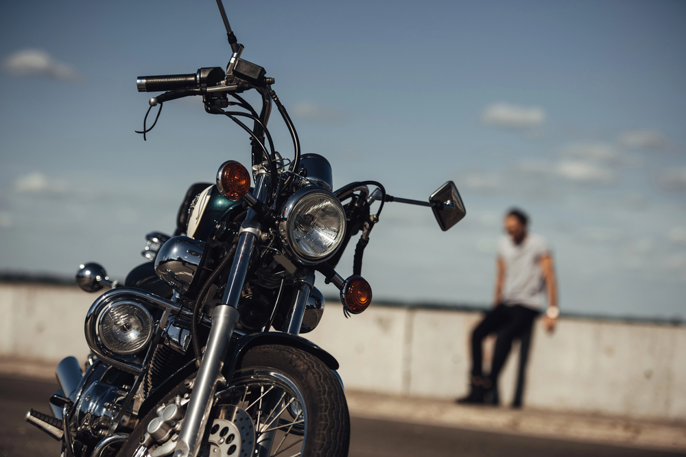 A close-up of a parked motorbike on a road, with a blurred person sitting on a concrete barrier in the background under a blue sky with scattered clouds.