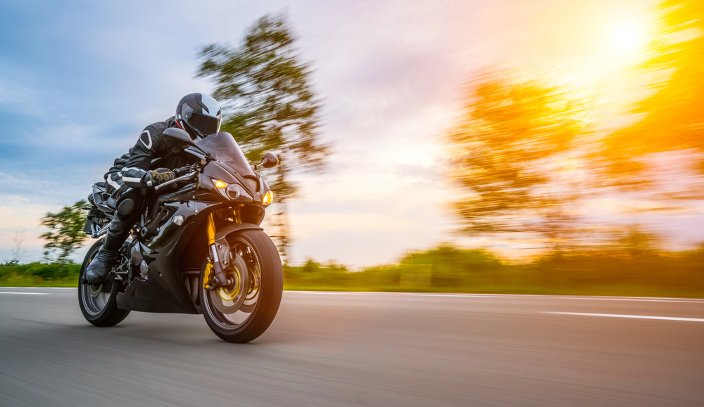 A person dressed in black riding gear speeds down a road on a black sports motorbike. The background is blurred with trees and sunlight, emphasising motion and speed.