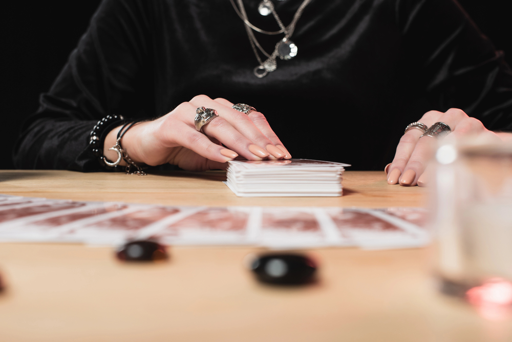 A person wearing rings and bracelets shuffles a pack of tarot cards on a wooden table, with more tarot cards laid out in front and small black stones nearby.