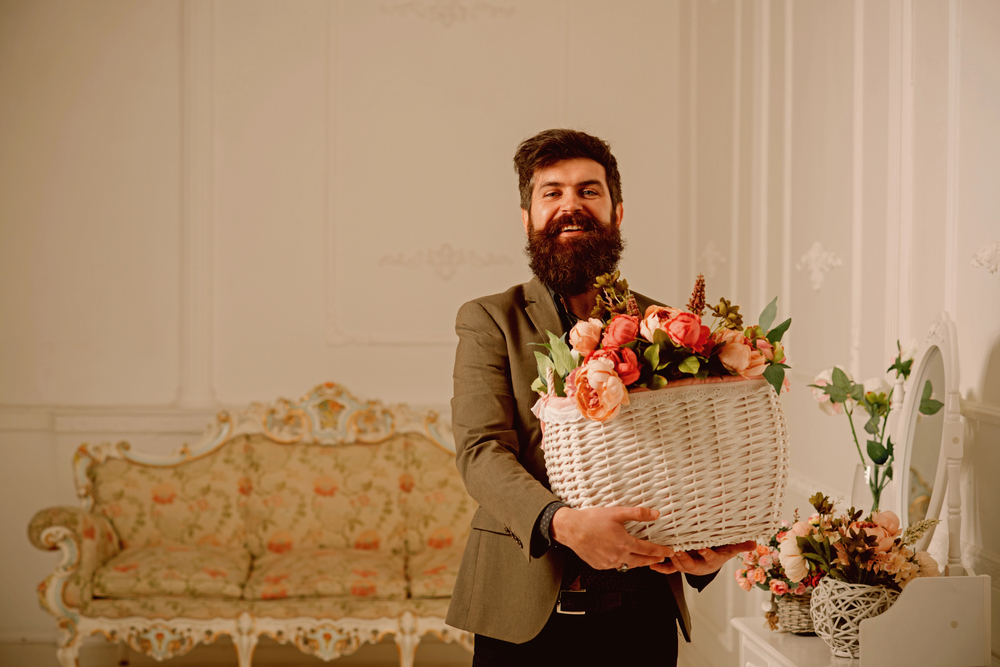 A bearded man in a blazer holds a large white wicker basket filled with colourful flowers, smiling in an elegant, vintage-style room with floral decor and ornate furniture.