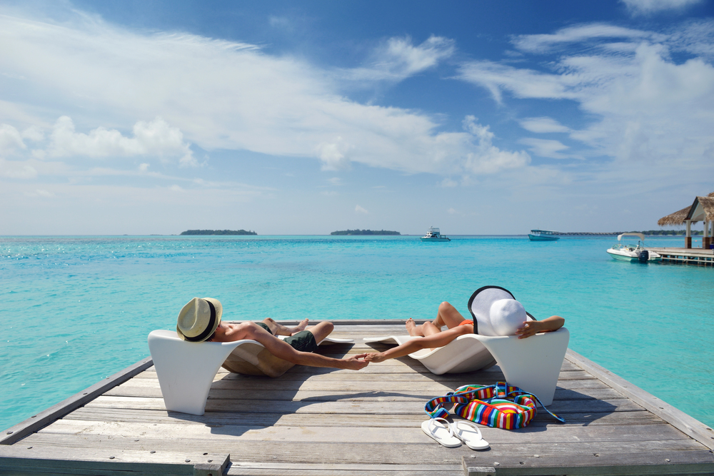 A couple relax on modern loungers on a wooden deck over turquoise water, holding hands, with hats and a colourful bag nearby under a blue sky with clouds. Boats and distant islands are visible in the background.