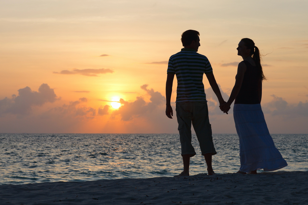 A couple stands on a beach holding hands and facing each other, silhouetted against a colourful sunset sky over the ocean.
