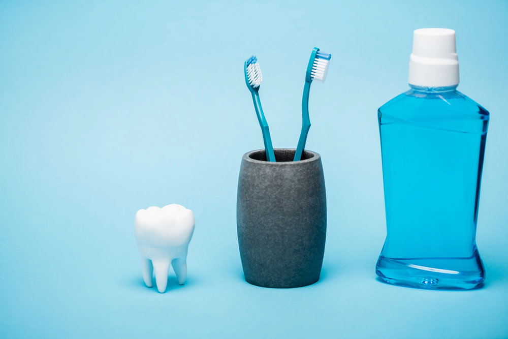 A model of a tooth, two blue toothbrushes in a grey cup, and a bottle of blue mouthwash are arranged on a light blue background.