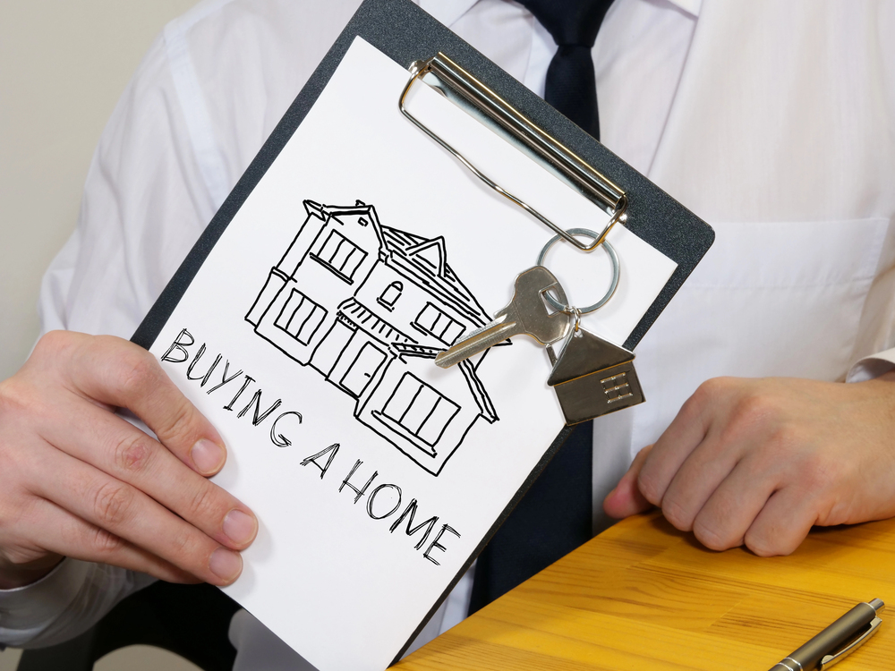 A person in a white shirt and tie holds a clipboard with a drawing of a house, a set of keys, and the words BUYING A HOME written below the illustration.