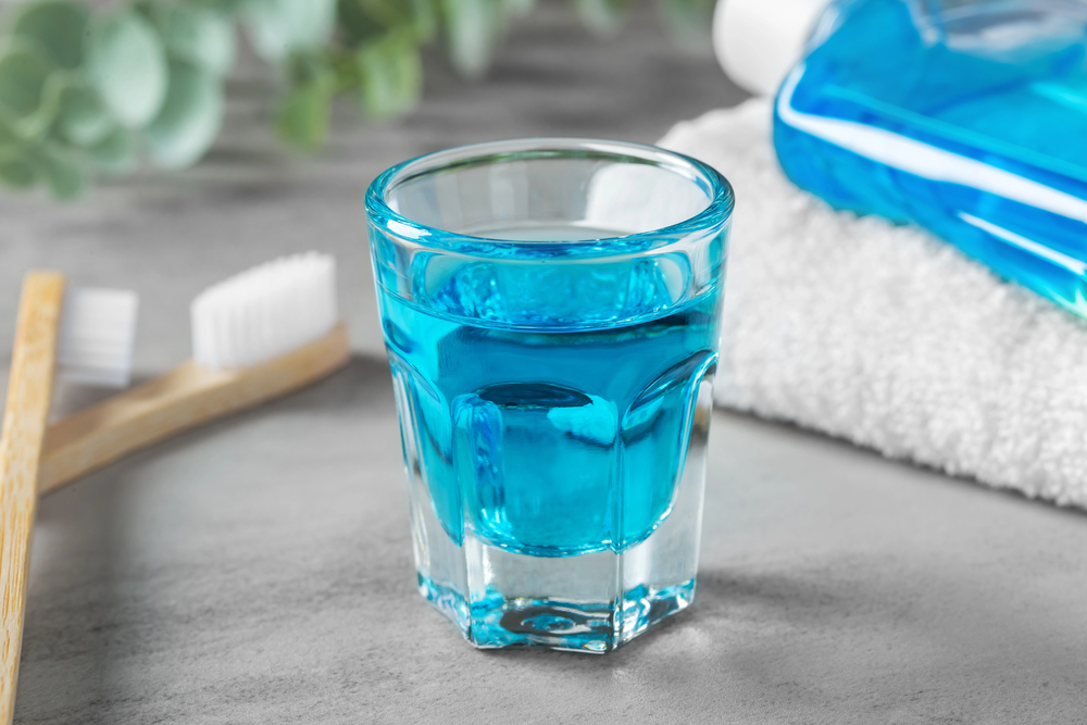 A glass filled with blue mouthwash sits on a grey surface next to a white towel, a toothbrush with white bristles, and green leaves in the background.
