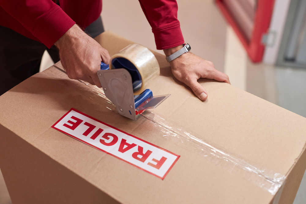 A person sealing a cardboard box with clear packing tape. The box has a large red and white FRAGILE label on top, indicating careful handling is needed.