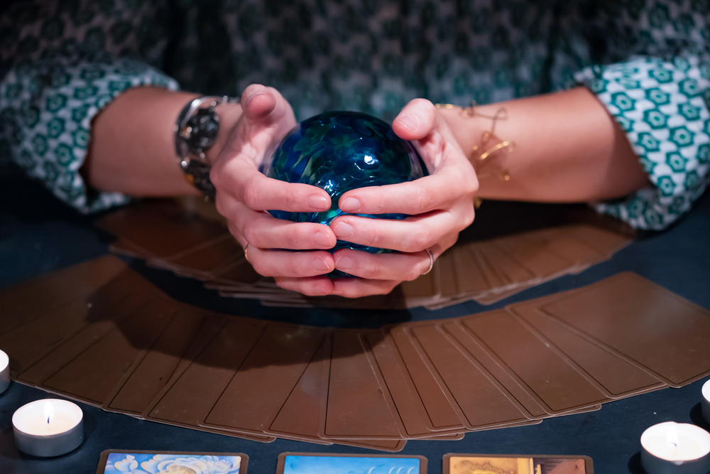 A person holding a blue crystal ball with both hands, surrounded by a spread of tarot cards and small lit candles on a table. The person is wearing a patterned shirt and bracelets.