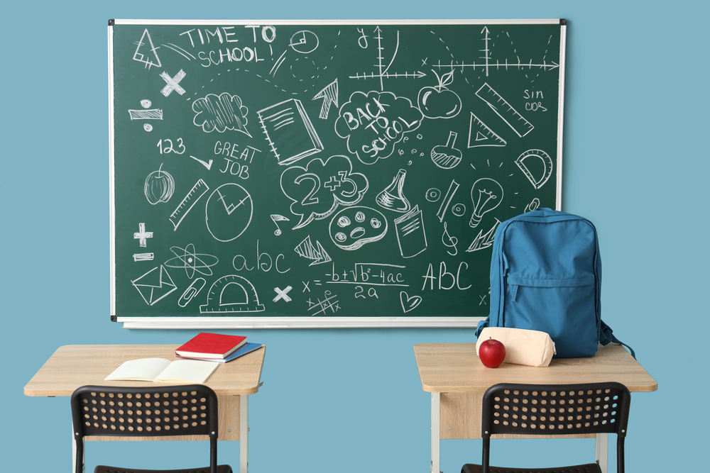 Two desks with notebooks, a rucksack, a lunch bag, and an apple in front of a chalkboard covered in school-related doodles, equations, and the words Back to School and Time to School on a blue wall background.