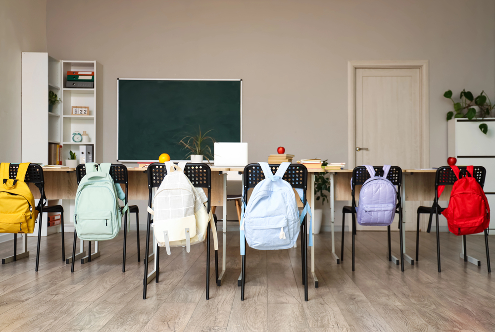 A classroom with a row of chairs, each with a colourful rucksack hanging on it, facing desks and a green blackboard. Books, apples, and plants are on the desks and shelves. The room is neat and well-lit.